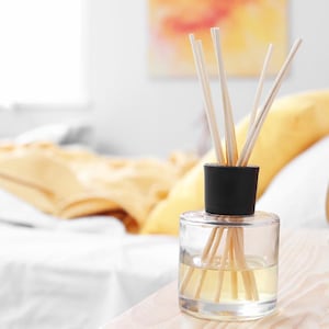 Puede incluir: Un difusor de aromaterapia de vidrio transparente con una tapa negra y varillas de madera. El difusor contiene un líquido amarillo claro y se encuentra sobre una superficie de madera. El fondo incluye una cama con almohadas amarillas.