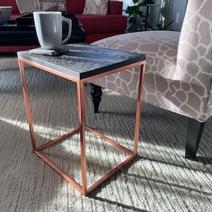 May include: A modern side table with a dark wood top and a copper-colored metal frame. The table is rectangular and has a minimalist design. A white mug sits on the table, resting on a gray coaster.