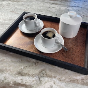 May include: A black wooden serving tray with a copper-colored interior holds two white coffee cups and saucers, a small spoon, and a white sugar bowl. The cups are filled with dark coffee.