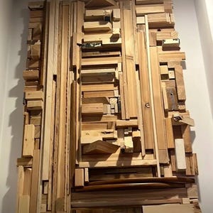 Puede incluir: Arte mural abstracto de madera compuesto por piezas de madera natural de varios tamaños y formas. La obra de arte está montada en una pared blanca, mostrando un diseño texturizado y tridimensional. Los tonos de madera varían de marrón claro a medio.