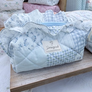 May include: A collection of quilted fabric bags in various sizes and patterns. The bags feature a mix of floral, gingham, and solid light blue fabrics. The main bag has a ruffled edge and a label that reads "Sewgud".
