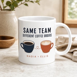 May include: A white ceramic mug featuring the dark blue text SAME TEAM DIFFERENT COFFEE ORDERS. Below the text are illustrations of a dark blue coffee cup and a terracotta coffee cup connected by a thin red heart line, with the names NADIA + ELLIS.