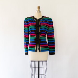 May include: A striped blazer with a black background and colorful stripes in pink, blue, green, and yellow. The blazer has black bows on the front.