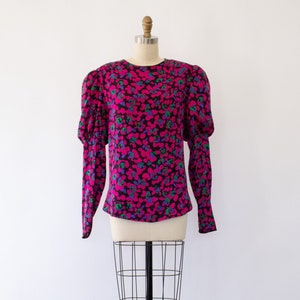 May include: A pink, black, green, and blue floral print long-sleeve blouse with puffed sleeves on a white background.
