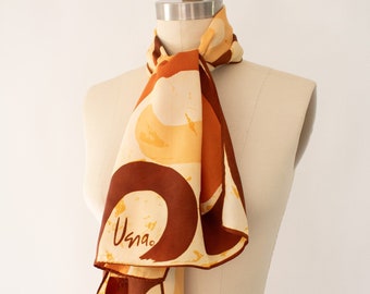 Può includere: Un foulard di seta marrone, giallo e bianco con un motivo geometrico e la parola "Usna" stampata su di esso. Il foulard è legato intorno al collo di un manichino.