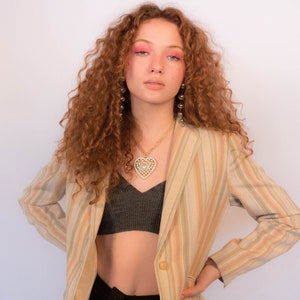 May include: A woman with long, curly red hair wearing a striped blazer, a black crop top, and a gold heart-shaped necklace. She is wearing gold earrings and has pink eyeshadow.