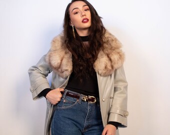 May include: A light grey leather coat with a faux fur collar. The coat is worn over a black top and blue jeans. A brown belt with a gold buckle is visible. The model is wearing gold earrings.