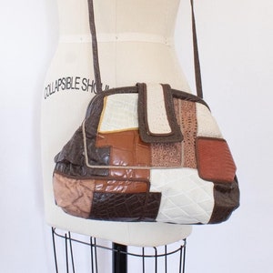 Puede incluir: Un bolso de hombro de cuero patchwork marrón, blanco y naranja con una correa larga. El bolso tiene un cierre de solapa y un diseño geométrico único.