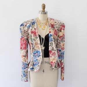 May include: A floral patterned blazer with a fitted silhouette and long sleeves. The blazer is in shades of cream, red, blue, and green. The blazer is on a white mannequin.