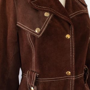 May include: Brown suede jacket with white stitching and gold buttons. The jacket features a notched collar, two front pockets, and a belt detail. A vintage-inspired piece.