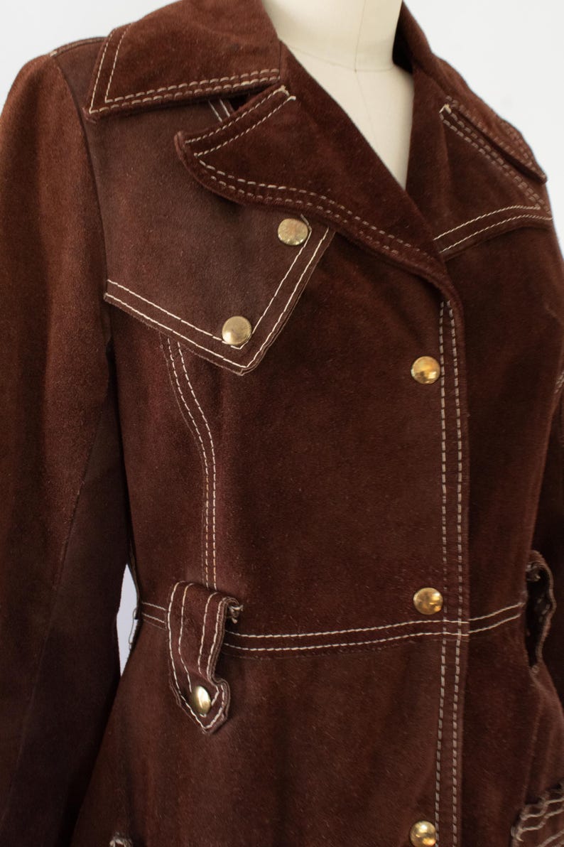 May include: Brown suede jacket with white stitching and gold buttons. The jacket features a notched collar, two front pockets, and a belt detail. A vintage-inspired piece.