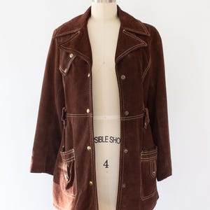 May include: A brown suede jacket with a notched collar, two front pockets, and gold-colored buttons. The jacket has white stitching details and is displayed on a mannequin. A vintage-style garment.