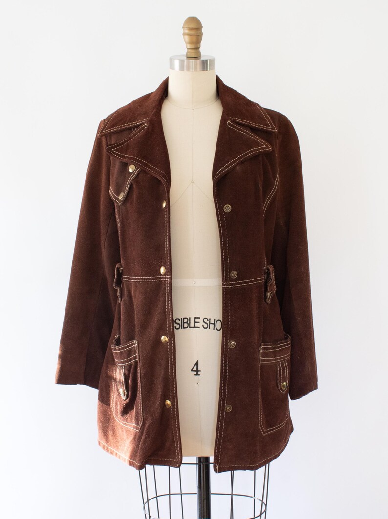 May include: A brown suede jacket with a notched collar, two front pockets, and gold-colored buttons. The jacket has white stitching details and is displayed on a mannequin. A vintage-style garment.