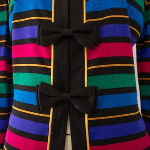 May include: A black jacket with a striped pattern in shades of pink, blue, green, and gold. The jacket has a black bow at the center of each stripe.