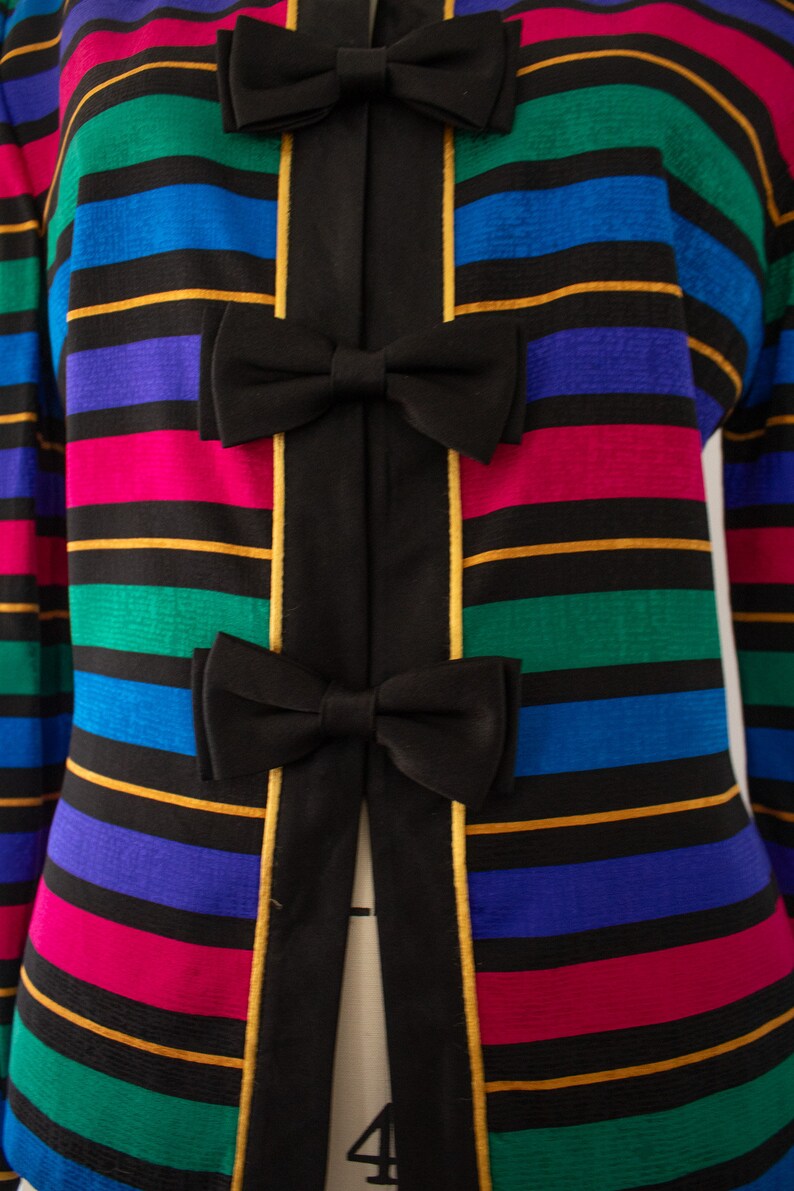 May include: A black jacket with a striped pattern in shades of pink, blue, green, and gold. The jacket has a black bow at the center of each stripe.