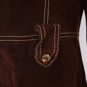 May include: Close-up of a dark brown garment with white stitching. The garment features a flap with a gold-colored button. The stitching is visible along the seams and around the flap, creating a contrasting detail.