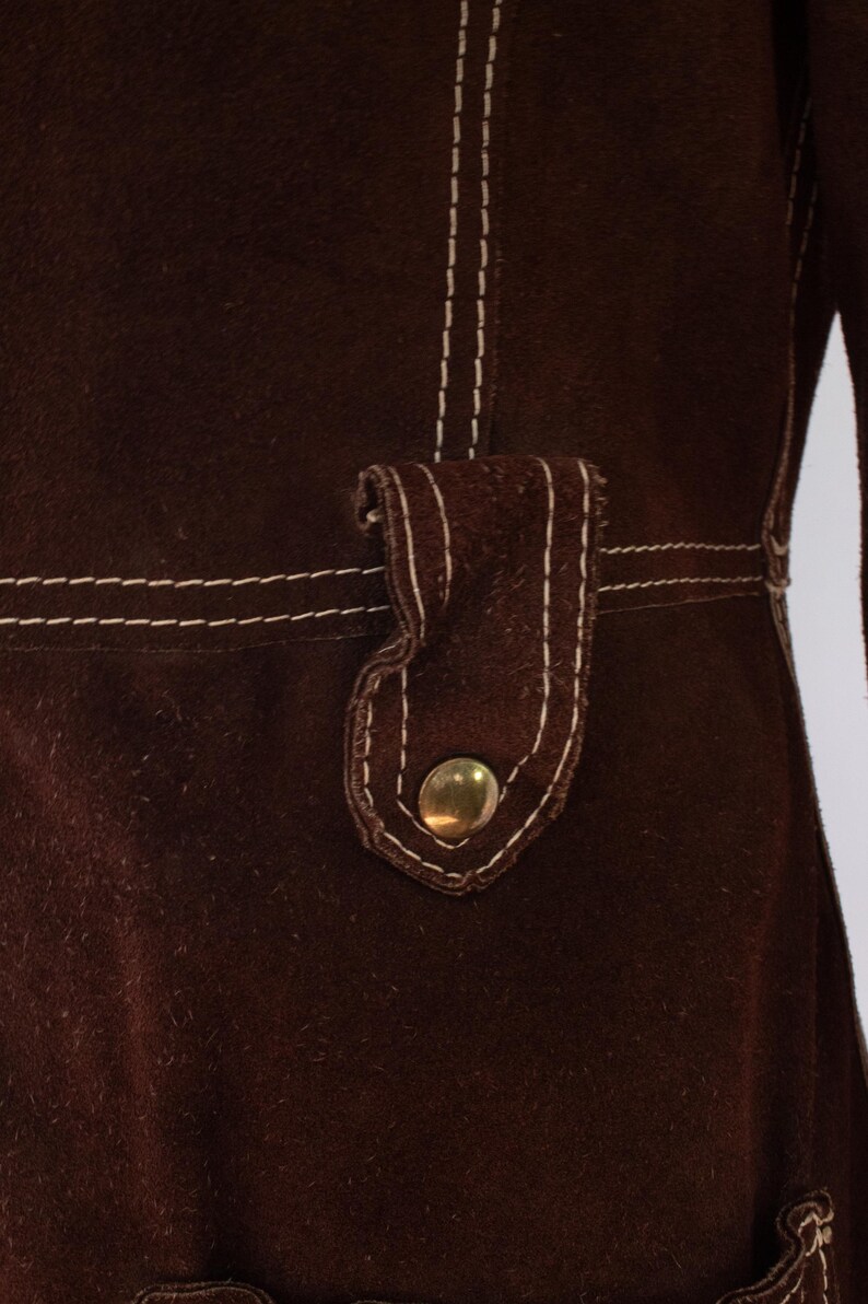 May include: Close-up of a dark brown garment with white stitching. The garment features a flap with a gold-colored button. The stitching is visible along the seams and around the flap, creating a contrasting detail.