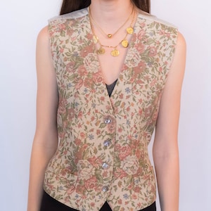 May include: A sleeveless floral patterned vest with a V-neckline and button closure. The vest is made of a beige fabric with a floral print in shades of pink, green, and blue.