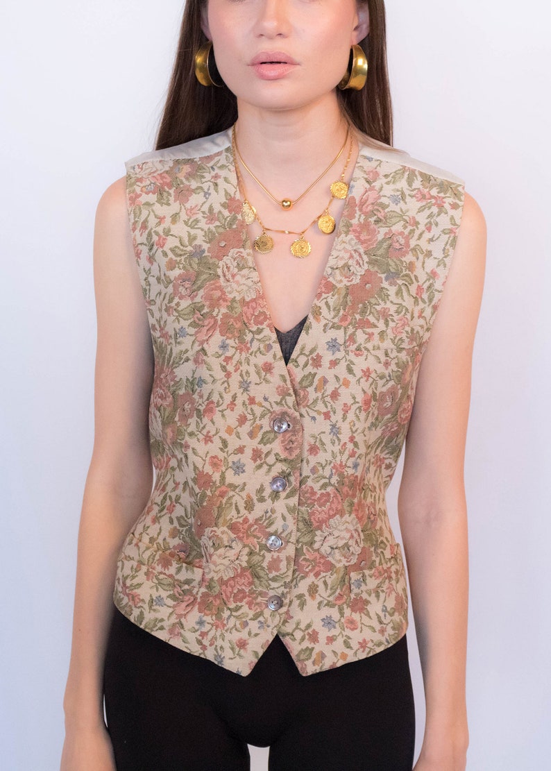 May include: A sleeveless floral patterned vest with a V-neckline and button closure. The vest is made of a beige fabric with a floral print in shades of pink, green, and blue.