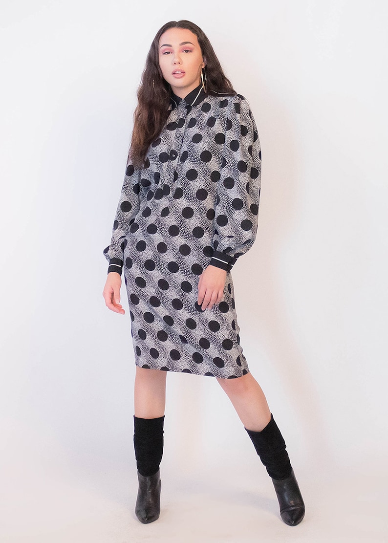 Peut inclure: Une robe &agrave; pois noirs et blancs avec un col et des manches longues. La robe a une silhouette ajust&eacute;e et arrive au niveau du genou. Le mannequin porte des bottes noires.