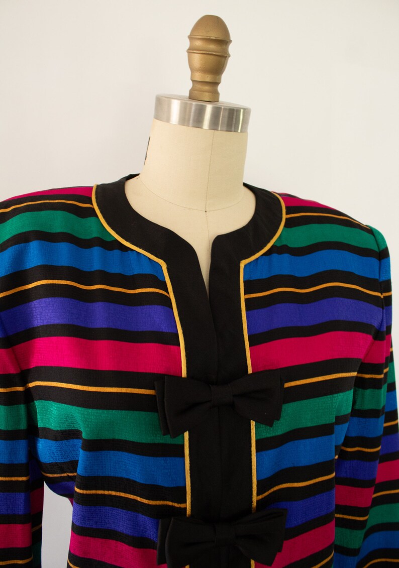 May include: A colorful striped blazer with a black collar and gold trim. The blazer has a V-neck and features a black bow at the front.