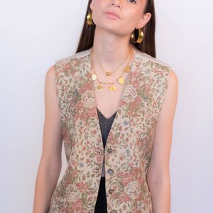 May include: A sleeveless floral patterned vest with a button closure. The vest is beige with pink, green, and blue flowers. The model is wearing a black top underneath the vest.