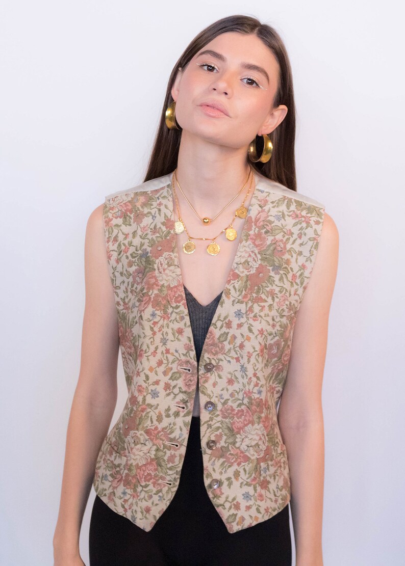 May include: A sleeveless floral patterned vest with a button closure. The vest is beige with pink, green, and blue flowers. The model is wearing a black top underneath the vest.