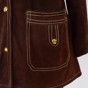 May include: A close-up of a brown suede jacket with white stitching and gold buttons. The jacket features a pocket with a flap and a button closure. The stitching is visible along the seams and around the pocket.