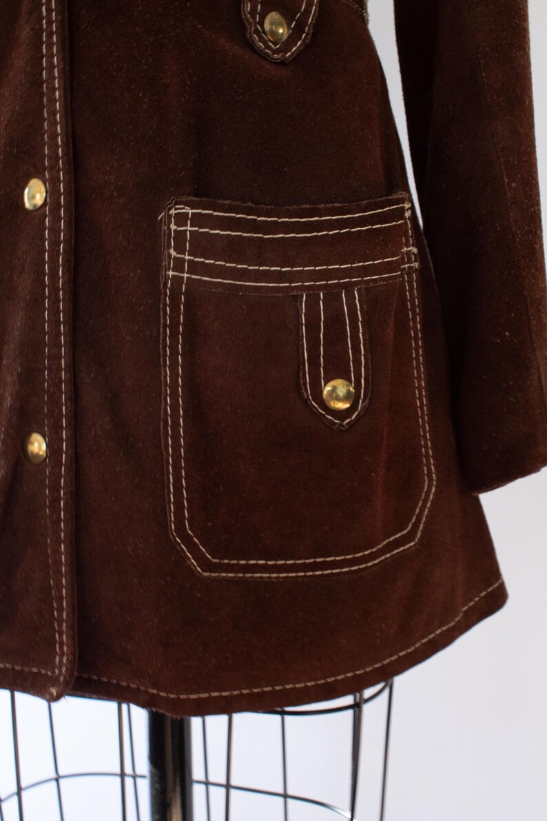 May include: A close-up of a brown suede jacket with white stitching and gold buttons. The jacket features a pocket with a flap and a button closure. The stitching is visible along the seams and around the pocket.