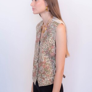 May include: A sleeveless floral patterned vest with a button closure. The vest is a light beige color with pink, green, and blue floral accents.