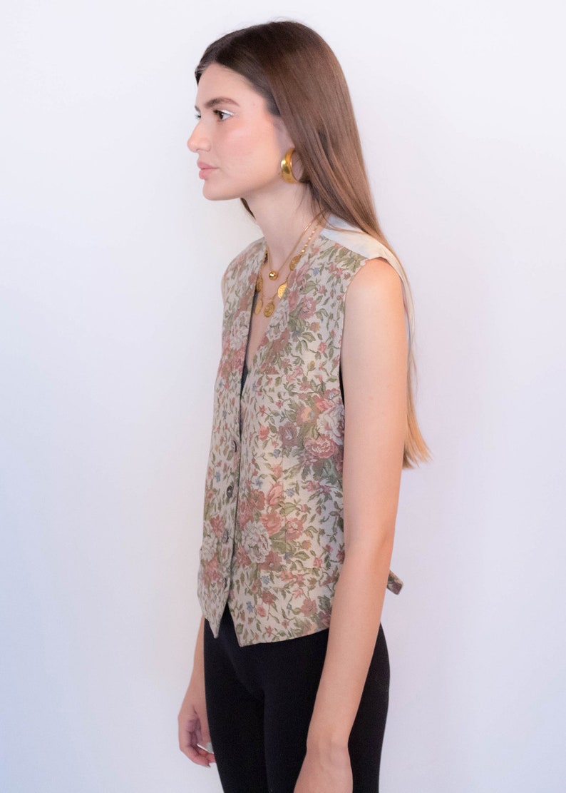 May include: A sleeveless floral patterned vest with a button closure. The vest is a light beige color with pink, green, and blue floral accents.