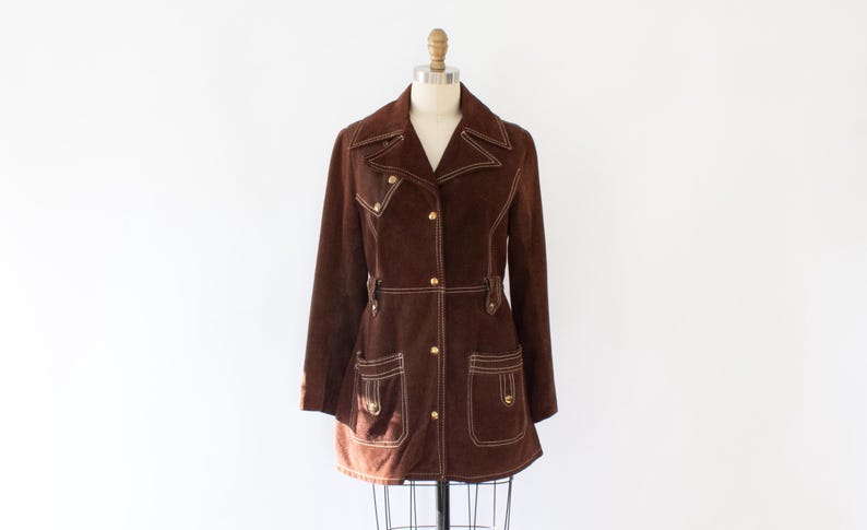 May include: A brown suede jacket with a notched collar, button closure, and front pockets. The jacket features white stitching and gold-colored buttons. It is displayed on a mannequin against a white background. A vintage-style coat.