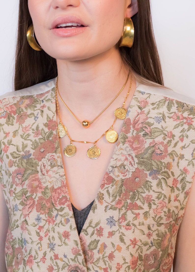 May include: A woman wearing a floral patterned vest with a gold chain necklace featuring a large pendant and a smaller chain with a gold ball pendant. She is also wearing gold hoop earrings.