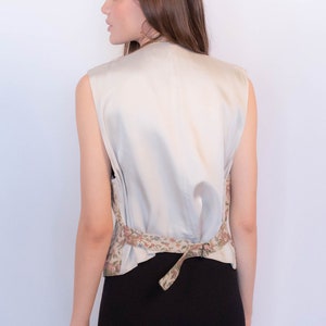May include: A sleeveless, cream-colored vest with a floral print around the waist. The vest is tied at the back with a matching fabric tie.