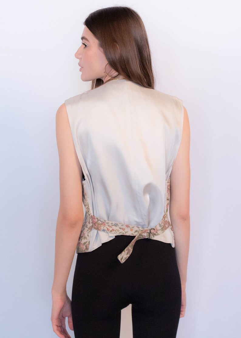 May include: A sleeveless, cream-colored vest with a floral print around the waist. The vest is tied at the back with a matching fabric tie.