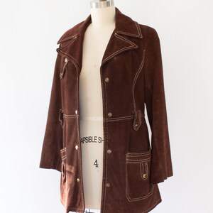 May include: A brown suede jacket with white stitching details. The jacket features a collared neckline, button closures, and two front pockets. The jacket is displayed on a mannequin.