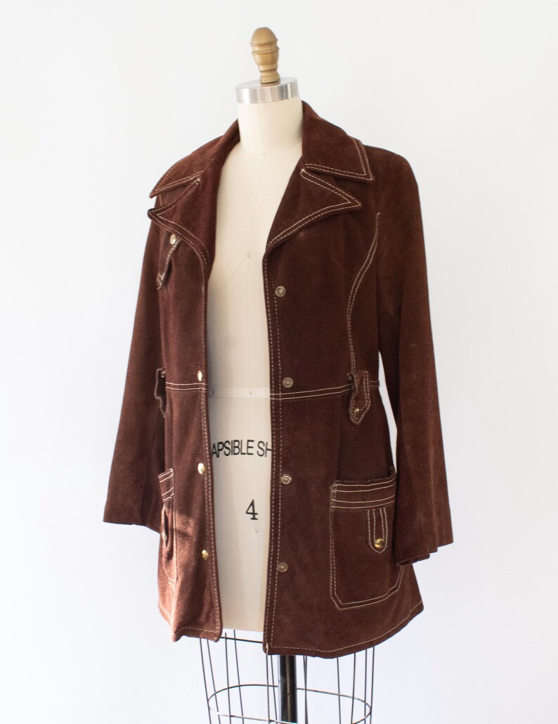May include: A brown suede jacket with white stitching details. The jacket features a collared neckline, button closures, and two front pockets. The jacket is displayed on a mannequin.