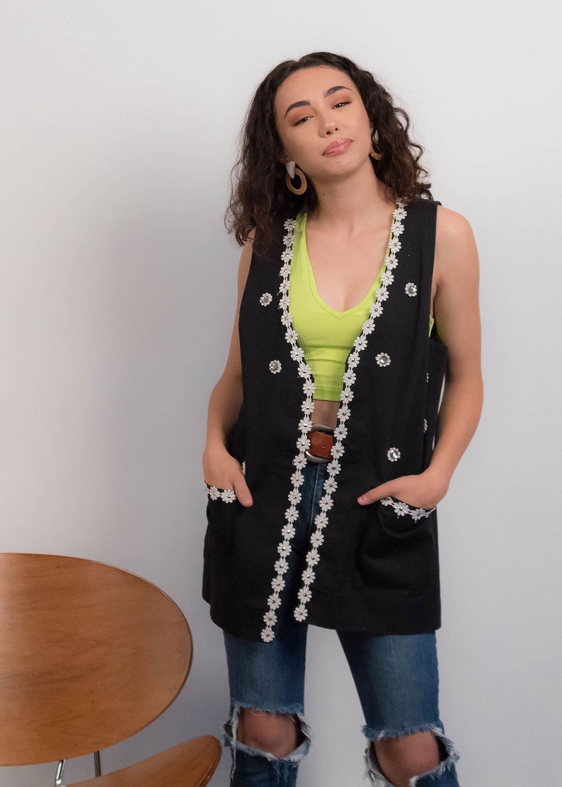 May include: A black sleeveless vest with white daisy-shaped trim and rhinestone accents. The vest has two pockets and is worn over a green tank top and blue jeans.