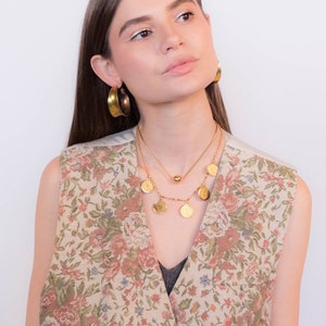 May include: A woman wearing a floral patterned vest with a gold chain necklace and gold hoop earrings.