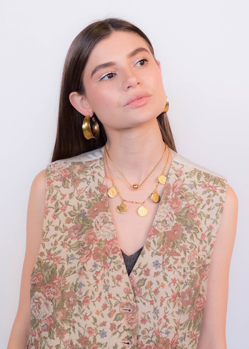 May include: A woman wearing a floral patterned vest with a gold chain necklace and gold hoop earrings.