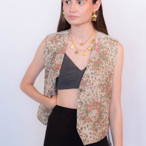 May include: A sleeveless floral patterned vest with a button closure. The vest is worn over a gray crop top and black pants.