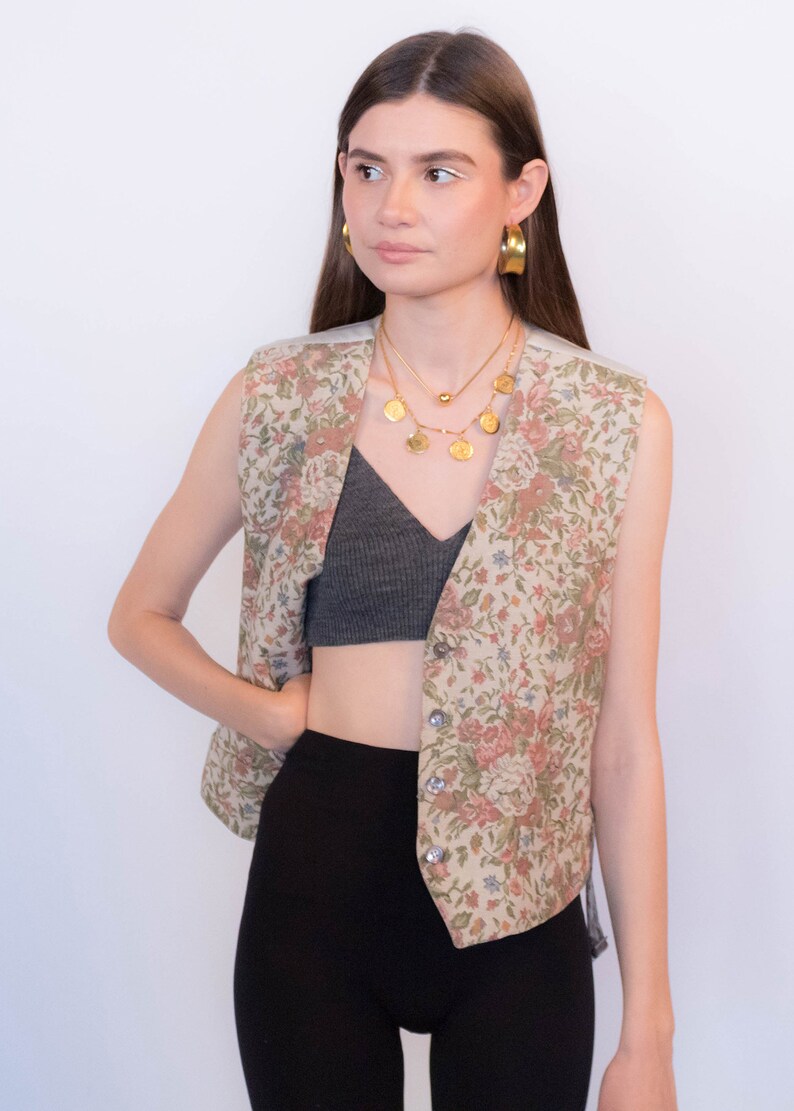 May include: A sleeveless floral patterned vest with a button closure. The vest is worn over a gray crop top and black pants.