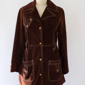 May include: A brown suede jacket with white stitching and gold buttons. The jacket has a notched collar, two front pockets, and a belt detail at the waist. The jacket is displayed on a mannequin.