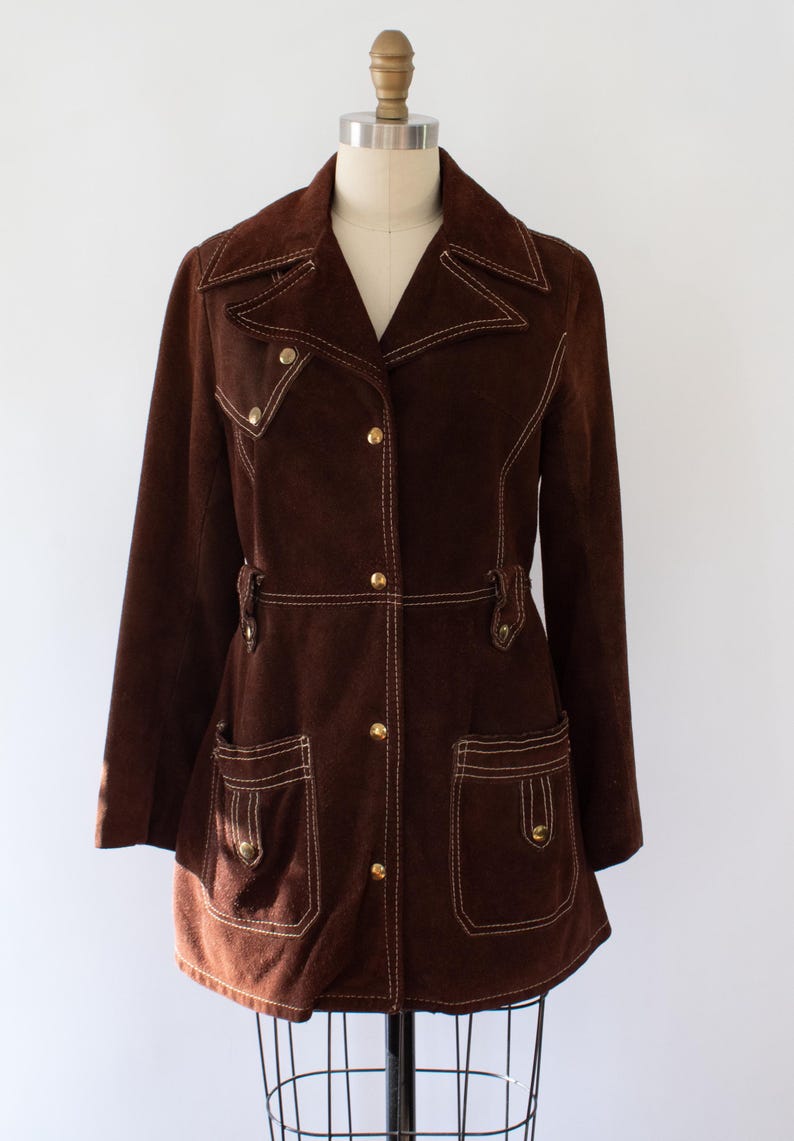 May include: A brown suede jacket with white stitching and gold buttons. The jacket has a notched collar, two front pockets, and a belt detail at the waist. The jacket is displayed on a mannequin.