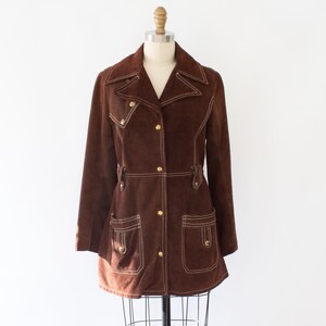 May include: A brown suede jacket with white stitching and gold buttons. The jacket has a notched collar, two front pockets, and a belt detail at the waist. The jacket is displayed on a mannequin.