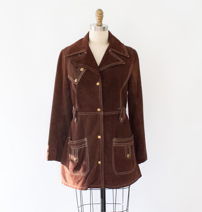 May include: A brown suede jacket with white stitching and gold buttons. The jacket has a notched collar, two front pockets, and a belt detail at the waist. The jacket is displayed on a mannequin.