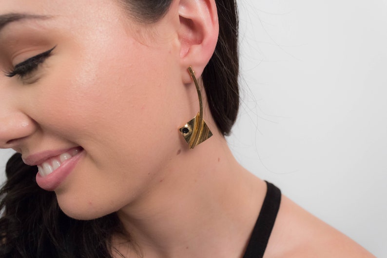May include: Gold tone geometric earrings with a black stone accent. The earrings are long and dangle.