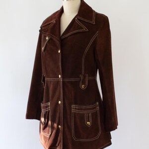 May include: A brown suede jacket with white stitching and gold buttons. The jacket has a notched collar, two front pockets, and a button closure. The jacket is displayed on a mannequin. The style is reminiscent of the 1970s.