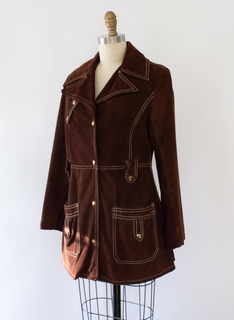 May include: A brown suede jacket with white stitching and gold buttons. The jacket has a notched collar, two front pockets, and a button closure. The jacket is displayed on a mannequin. The style is reminiscent of the 1970s.