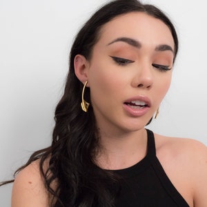 May include: A woman with long dark hair wearing gold hoop earrings. She is wearing a black tank top and has a neutral makeup look with winged eyeliner.